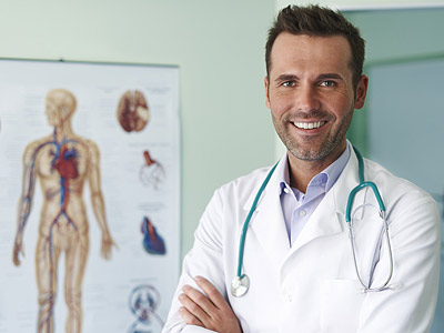 Ein Arzt lächelt freundlich mit einem Stethoskop um den Hals, vor einer Wandtafel mit menschlicher Anatomie.
